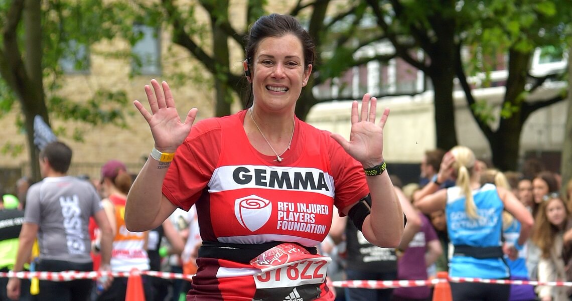 Woman called Gemma holding both hands in the air as she runs past the camera during a running event. She is wearing an Injured Players Foundation vest with red and white horizontal stripes.